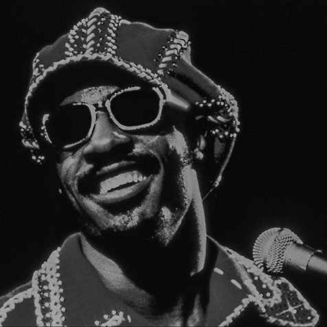 Stevie Wonder souriant, avec ses lunettes et un chapeau, devant un microphone